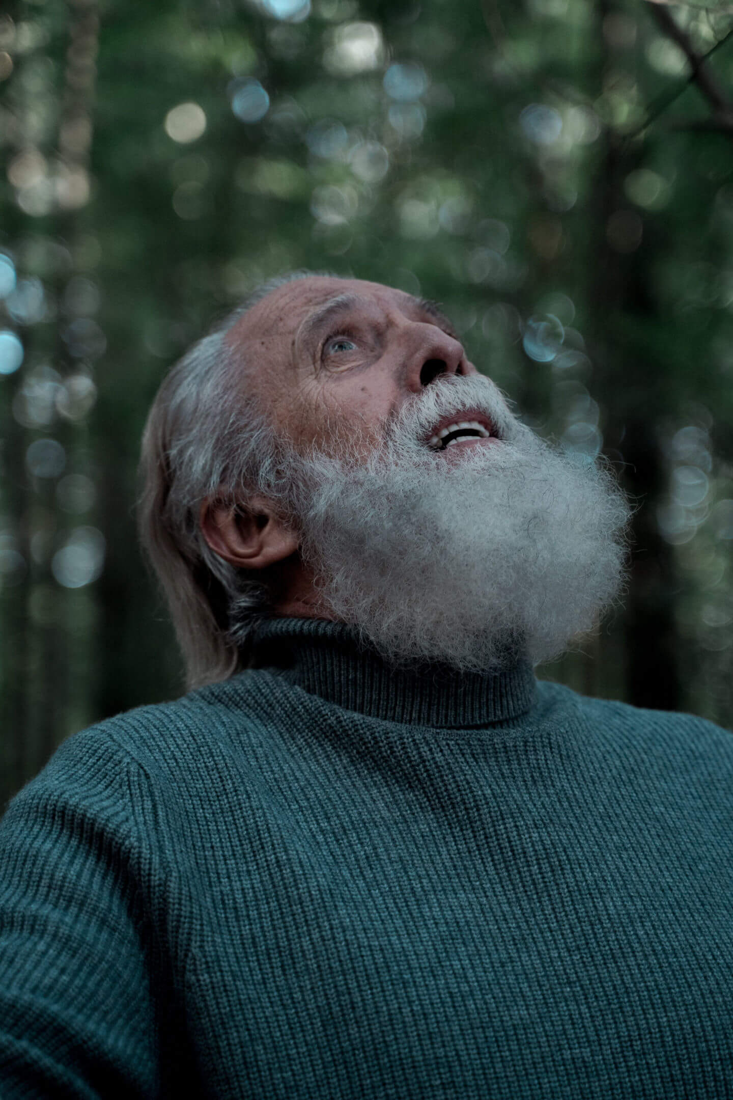 Older man standing in the woods, looking up at the sky flustered, or bewildered.
