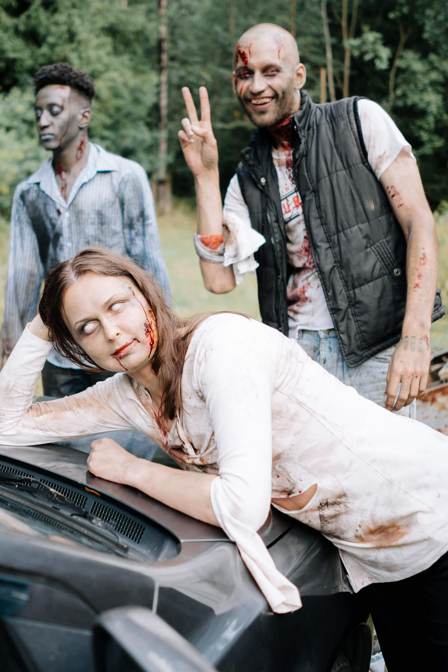 A group of zombies posing for a photo. One is leaning on a car, another behind is flashing a peace sign, while a 3rd looks on in the background.