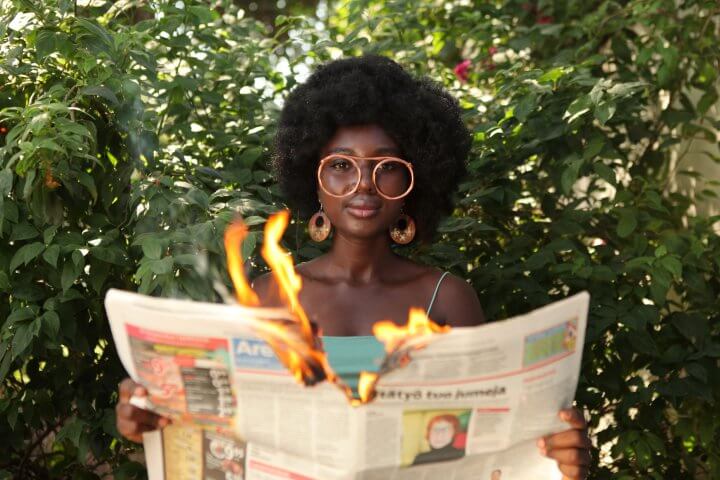 Woman with glasses facing the camera, holding a newspaper that is on fire and being torn in half