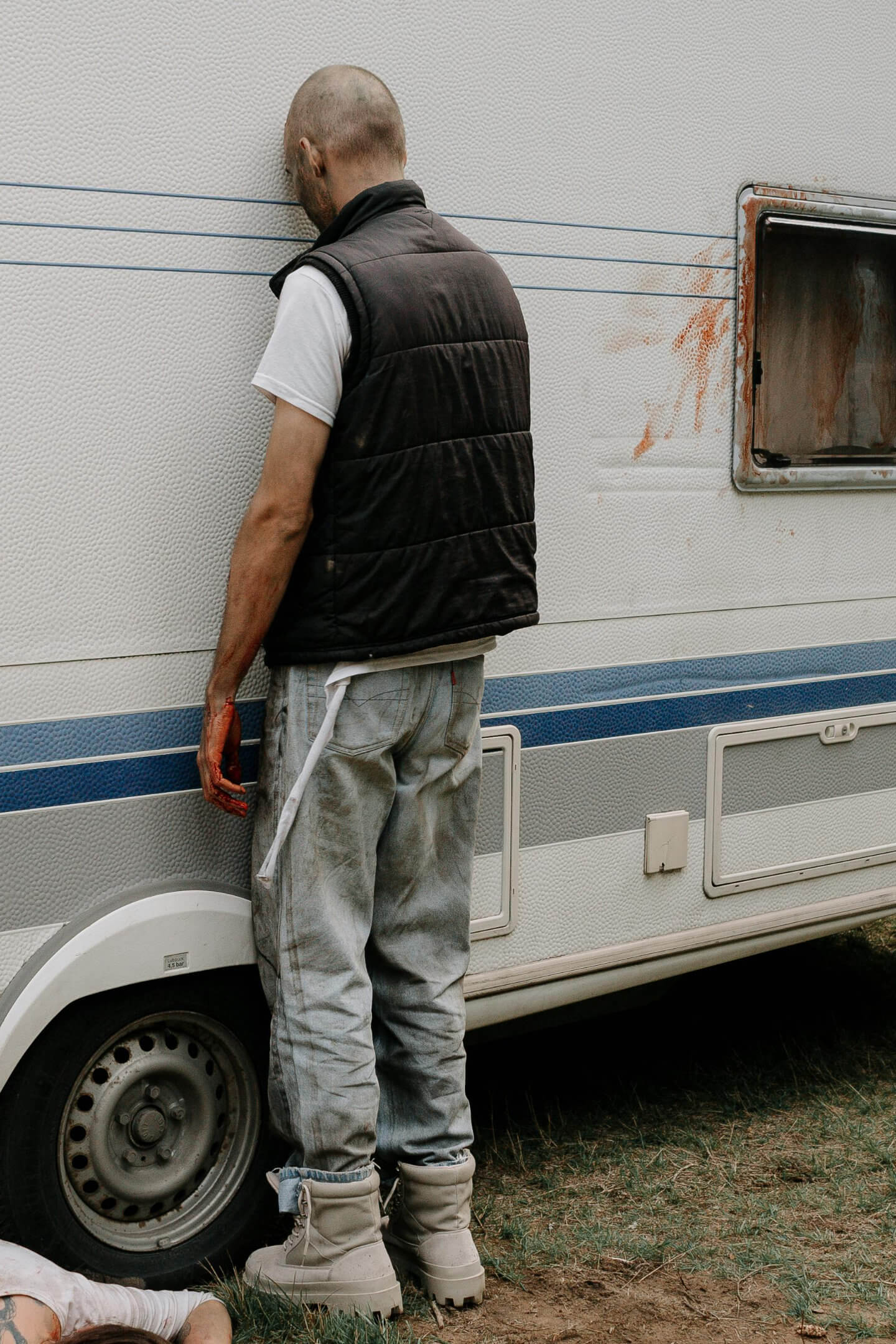 Undead man in grey jeans and boots with a short sleeve white t-shirt and black vest stands facing a mobile home exterior wall, leaning his head against the mobile home exterior wall.